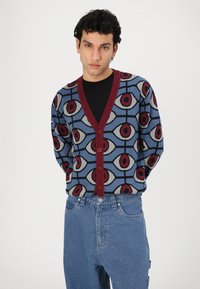 Cardigan cu un model albastru și margine roșie, având un motiv cu ochi și nasturi pe față. Purtați peste o cămașă neagră și asortați cu blugi.
