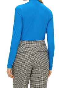 Langarmige blauwe top met een hoge kraag, gecombineerd met zwart-witte houndstooth-patroonbroek. Gladde stoftextuur zichtbaar.