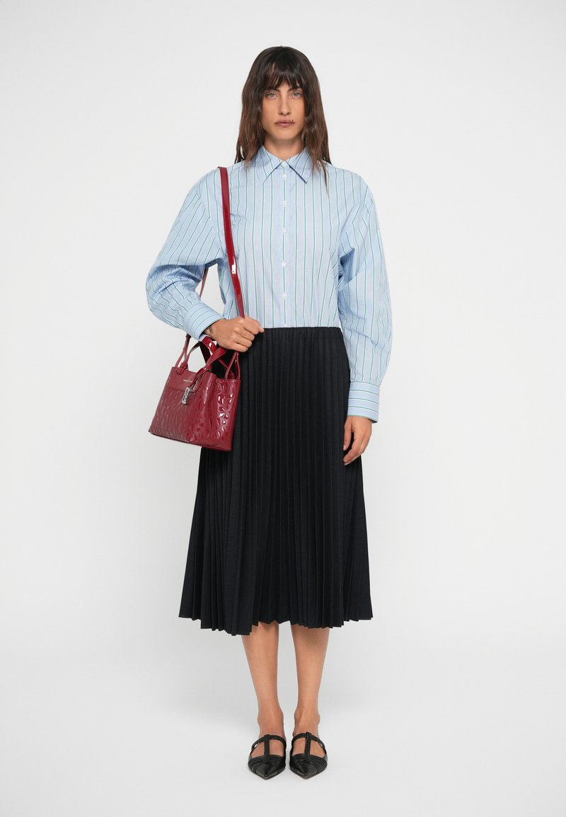 Chemise à rayures bleues et blanches avec des manches bouffantes, associée à une jupe plissée noire. Sac à main en cuir synthétique rouge, chaussures plates noires.