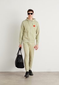 Hoodie verde chiaro con zip e pantaloni abbinati, dotati di una piccola toppa rossa, abbinati a sneakers nere e a una borsa nera. Tessuto liscio, vestibilità rilassata.