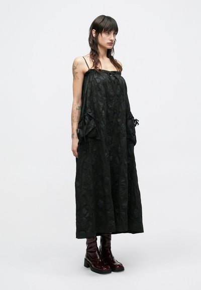 Μαύρο αμάνικο μάξι φόρεμα με υφασμένο φύλλωμα, πλαϊνές τσέπες και ρυθμιζόμενους δεσμούς στη μέση, σε συνδυασμό με γυαλιστερά μπορντό μποτάκια αστραγάλου.