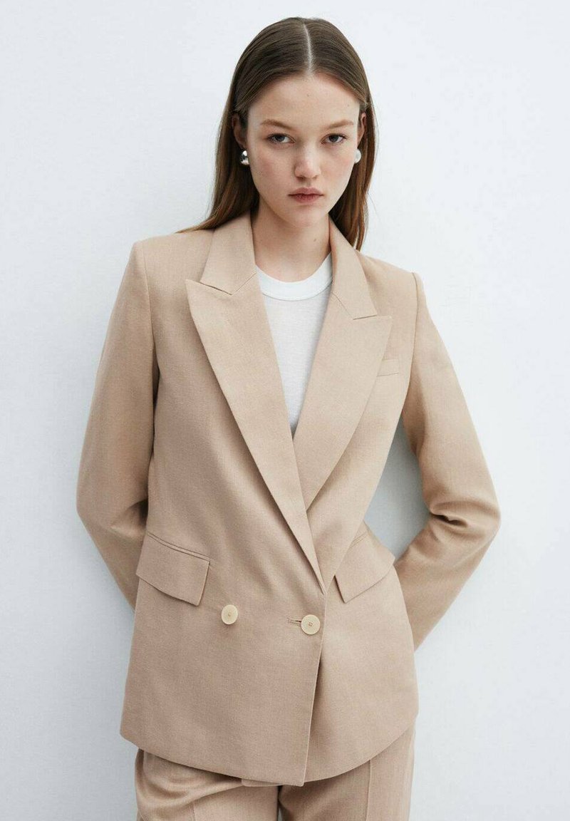 Blazer beige su misura con design doppiopetto, rever a punta, due tasche frontali e tessuto liscio. Indossato sopra un top bianco.