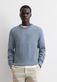 Hellblauer Strickpullover mit strukturiertem Muster, geripptem Kragen und langen Ärmeln, über einem weißen Hemd getragen und mit beigefarbenen Hosen kombiniert.