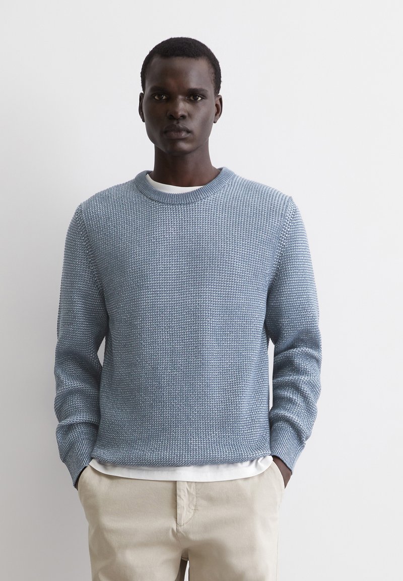 Hellblauer Strickpullover mit strukturiertem Muster, geripptem Kragen und langen Ärmeln, über einem weißen Hemd getragen und mit beigefarbenen Hosen kombiniert.