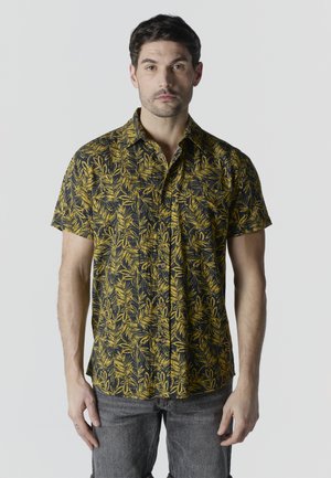 Joven con camisa negra de manga corta con patrones de hojas amarillas y jeans grises, de pie contra un fondo claro y sencillo.