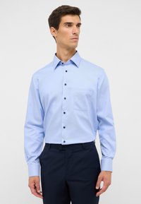 Camicia azzurra chiara a maniche lunghe con colletto button-down, dotata di una tasca sul petto e bottoni scuri su un tessuto liscio.