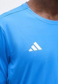 Άνδρας που φοράει φωτεινό μπλε αθλητικό μπλουζάκι με λευκό λογότυπο Adidas με τρεις ρίγες στο στήθος, ορατό από τον λαιμό έως το μέσο του κορμού.