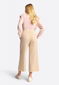 Pantalons beige à jambes larges avec une coupe ajustée, dotés de poches arrière. Associés à un top à manches longues rose clair et des chaussures claires.