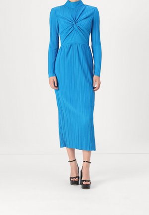Femme portant une robe midi plissée bleu vif à manches longues avec un détail de nœud torsadé au niveau de la poitrine et des sandales noires à talons hauts.