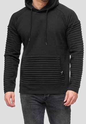 Man wearing a black hoodie with ribbed textured sleeves and a ribbed front pocket, paired with dark grey jeans.