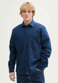 Koszula dżinsowa w ciemnoniebieskim kolorze z zapięciem na guziki, dwiema kieszeniami na piersi, długimi rękawami i gładką fakturą. Posiada czarne guziki i kołnierz.