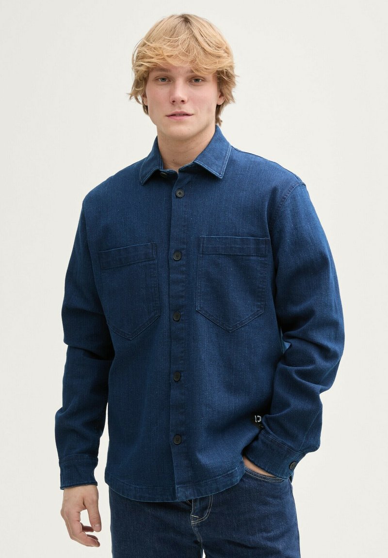 Koszula dżinsowa w ciemnoniebieskim kolorze z zapięciem na guziki, dwiema kieszeniami na piersi, długimi rękawami i gładką fakturą. Posiada czarne guziki i kołnierz.