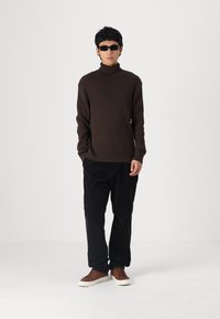 Pulover cu guler înalt cafeniu, pantaloni largi negri și adidași cafenii cu talpă albă. Model purtând ochelari de soare închiși la culoare, stând în fața unui fundal alb.