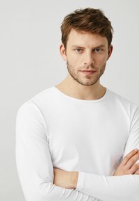 Camiseta blanca de manga larga hecha de una tela suave, con cuello redondo y un diseño ajustado, llevada por una persona con los brazos cruzados.