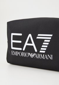 Juodas audinio maišelis su dideliu baltais spaudiniais "EA7 EMPORIO ARMANI" ir dryžuotais akcentais. Stačiakampio formos, su lygiu paviršiumi.