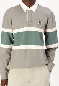 Mężczyzna w koszulce rugby z długim rękawem w poziome paski w odcieniach szarości, bieli i zieleni, z małym znaczkiem na piersi, w zestawie z beżowymi spodniami.