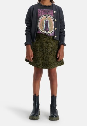 Ragazza con cardigan nero a bottoni, t-shirt grafica nera, gonna verde con stampa leopardata e stivali neri chunky, in piedi contro uno sfondo bianco.