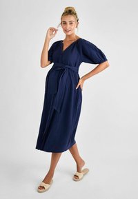 Abito midi blu navy con bottoni, maniche a sbuffo, vita con legatura e tessuto testurizzato. Indossato con sandali a scivolo beige annodati.