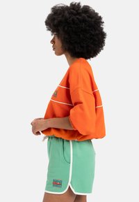 Sweatshirt oversized portocalie cu dungi albe și un logo grafic. Asortat cu pantaloni scurți verzi cu margini albe și un patch cu logo.