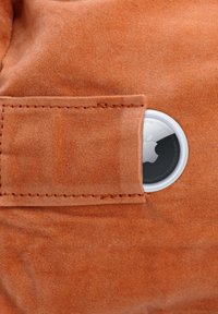 Apple AirTag teilweise sichtbar in einer braunen Wildledertasche mit sichtbaren Nähten an der Taschenklappe.