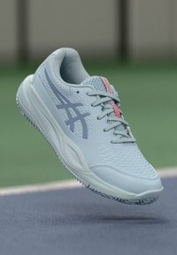 Hellblaue Asics-Laufschuhe mit grauem Logo und pinkem Zungendetail, über der Sportplatzoberfläche schwebend.