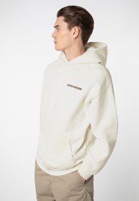 Cremefarbener Hoodie mit einer Kängurutasche, ausgestattet mit dem Schriftzug "WORLDWIDE" in brauner Schrift, aus weichem Baumwollmaterial. Lässige Passform mit Kapuze.