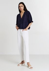 Donna che indossa una blusa blu navy a V con maniche a puff, pantaloni bianchi a gamba dritta e scarpe con tacco a kitten open-toe bianche, con le mani nelle tasche, su uno sfondo neutro.