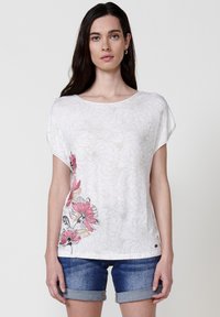 Haut floral à manches courtes blanches avec des contours de fleurs dans des teintes claires ; présente des accents floraux colorés sur le côté gauche. Texture de tissu douce.