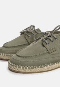 Chaussures en toile vert olive avec un design à lacets, bout rond, et détails en corde de jute le long de la semelle extérieure. Tissu texturé et œillets en métal.