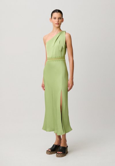 Donna in piedi che indossa un abito verde chiaro monospalla con uno spacco e sandali neri con zeppa e suola intrecciata, su uno sfondo chiaro e semplice.