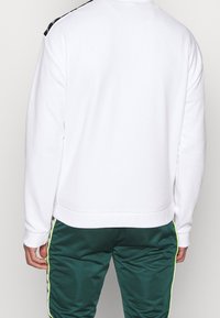 Bílá mikina s černým detailem na ramenou, se zaobleným výstřihem a žebrovanými lemy na rukávech. Kombinována se zelenými saténovými tepláky s neonovými akcenty.