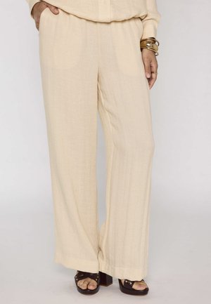 Pantaloni beige a gamba larga abbinati a sandali con tacco marrone scuro e braccialetti dorati al polso destro.