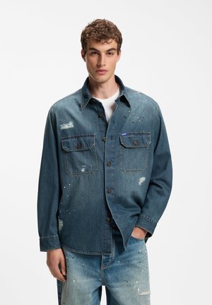 Ung mand iført en blå denimjakke og matchende jeans med slidte lapper og sprøjtede malingsdetaljer, stående mod en ensfarvet baggrund.