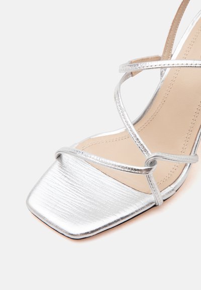 Sandale métallisée argentée à brides avec bout carré et semelle intérieure beige sur fond clair uni.
