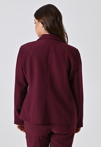 Frau mit langen braunen Haaren, die ein dunkelviolettes Blazer und passende Hosen trägt, stehend und mit dem Rücken zu einer schlichten hellen Hintergrund.