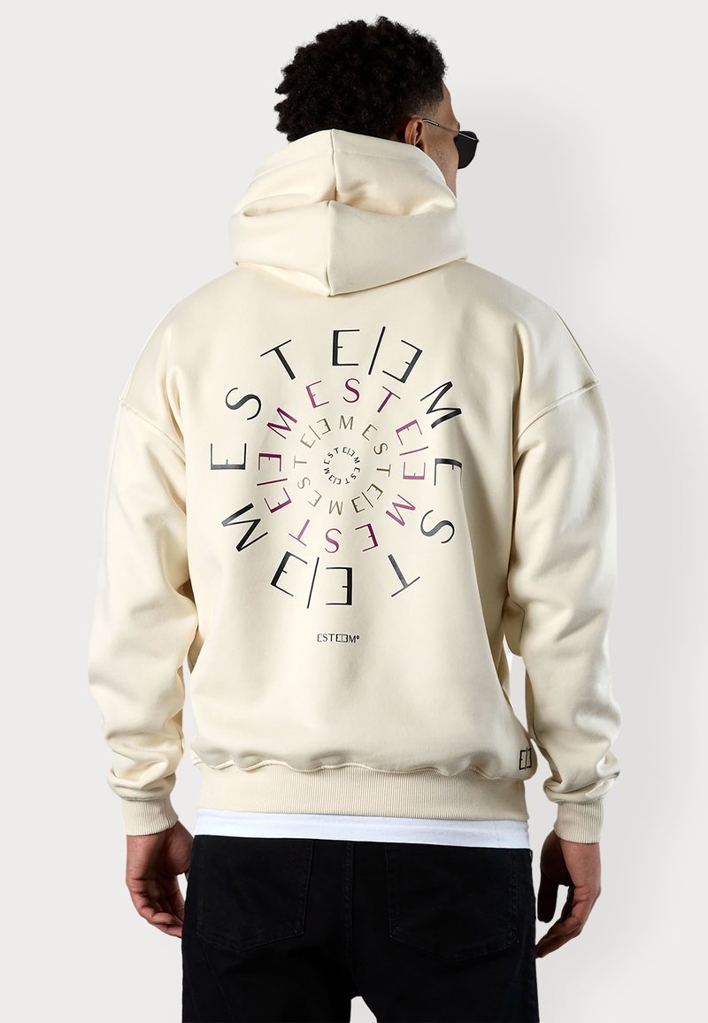 Homme portant un sweat à capuche crème avec un texte circulaire noir et rouge "ESTEEM" dans le dos, associé à un pantalon noir et des lunettes de soleil, de dos.
