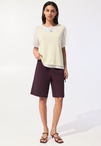 Canottiera giallo chiaro sopra una maglietta bianca, abbinata a pantaloni corti viola scuro. Tessuti lisci e vestibilità casual. Accessori minimi includono una collana.