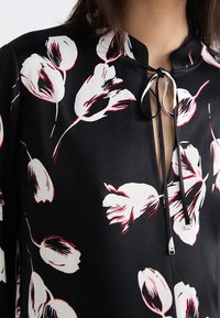 Blouse fleurie noire ornée de motifs de tulipes blanches et rouges, avec un petit col et une fermeture avant à nouer avec des cordons noirs et des accents métalliques.