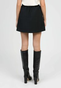 Femme se tenant de dos, portant une jupe noire et des bottes en cuir à talons noirs jusqu'aux genoux, sur un fond uni.