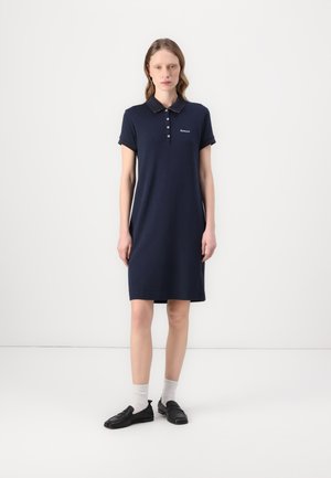 Granatowa sukienka polo z krótkimi rękawami, z trzema guzikami i detalem logo. Gładki materiał bawełniany, do kolan, zestawiona z czarnymi loafersami i jasnymi skarpetkami.