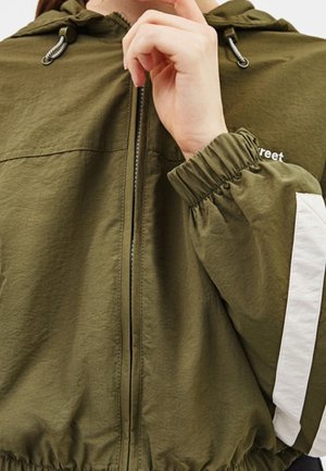 Person trägt eine olivgrüne Jacke mit Reißverschluss, elastischen Bündchen und weißem Streifen am Ärmel, Hand nahe am Gesicht erhoben.