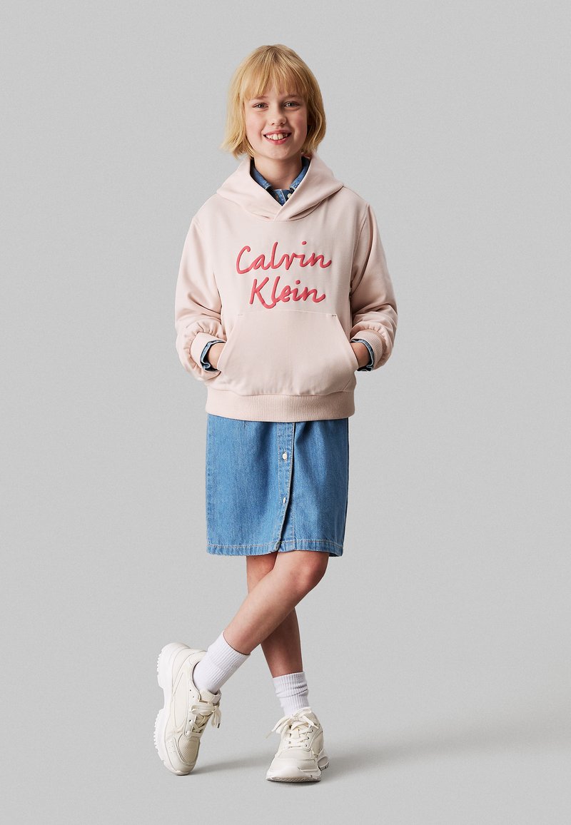 Felpa rosa con logo "Calvin Klein", tasca frontale, indossata sopra una gonna di denim. Il modello indossa sneaker bianche e calzini alla caviglia.