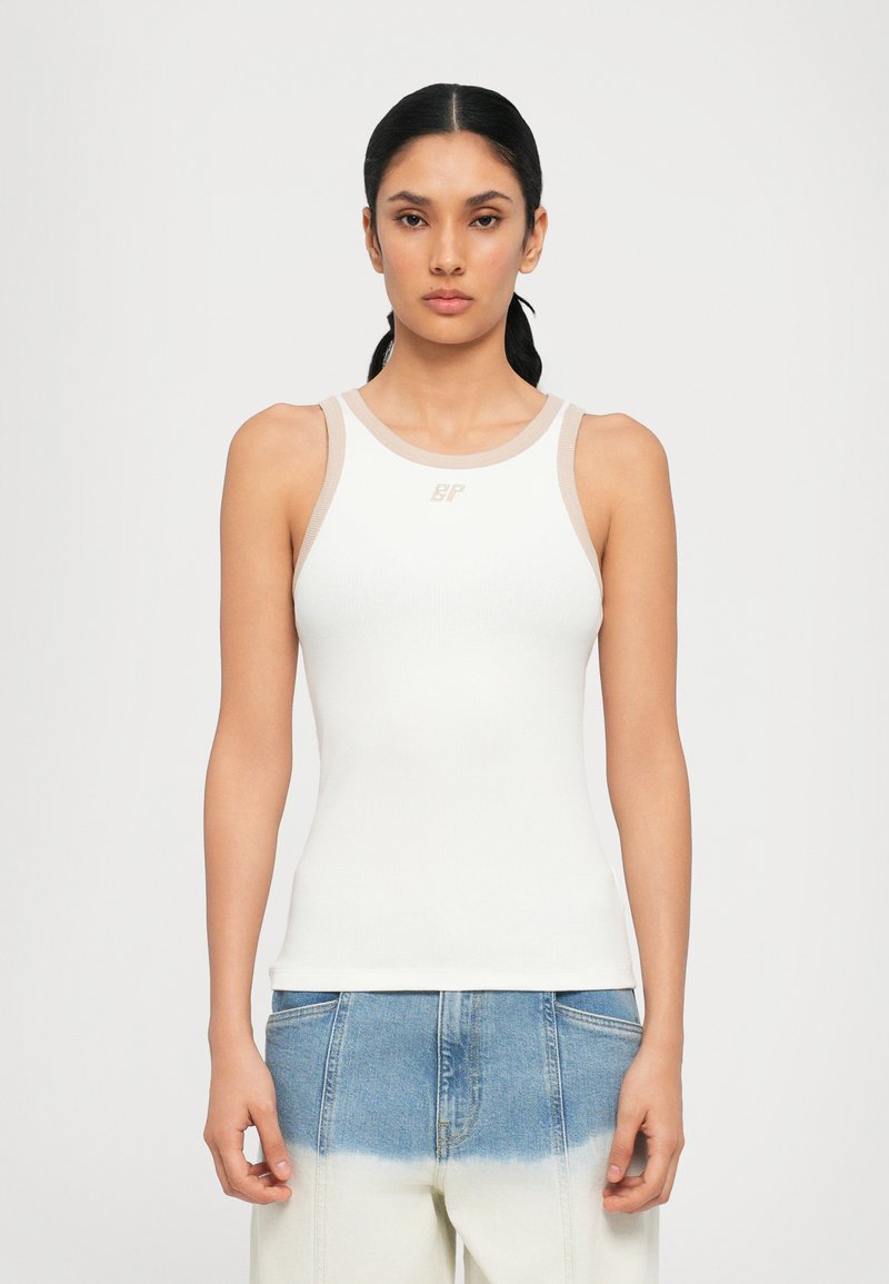 White ribbed tank top with a round neckline and light beige trim; features a subtle logo at the center. Paired with blue gradient denim pants.