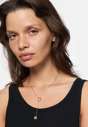 Goudkleurige ketting met een ronde hanger en een schakel, gedragen over een zwarte ribbed tanktop. Gladde textuur en eenvoudig ontwerp.
