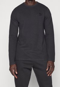 Schwarzes langärmliges Baumwoll-T-Shirt mit strukturiertem Muster, Rundhalsausschnitt und gesticktem Puma-Logo auf der Brust. Kombiniert mit schwarzen Hosen.
