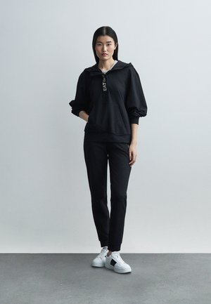 Žena stojaca v čiernej mikine s kapucňou a nohaviciach, s bielymi teniskami, na pozadí jednofarebnej šedej steny.