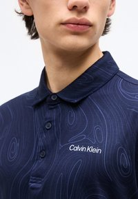 Gros plan d'une personne portant un polo bleu marine avec des motifs abstraits en spirale et un logo Calvin Klein blanc visible sur la poitrine.