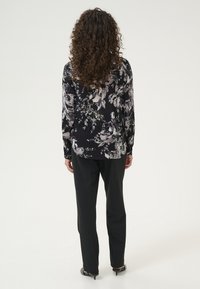 Blouse fleurie à manches longues noire avec des motifs blancs et gris, associée à un pantalon noir ; présente un ourlet arrondi et des poignets boutonnés.