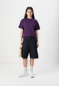 Purple short-sleeve t-shirt with a small logo, paired with dark denim shorts that reach the knees, white high-top sneakers, and a beige bag.