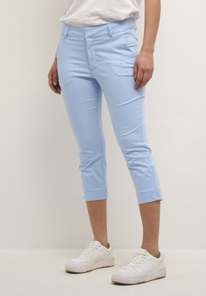 Person iført lyseblå afkortede bukser og hvide sneakers, stående mod en ensfarvet neutral baggrund.
